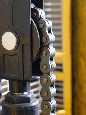 Close-up of a heavy-duty roller chain engaged in a black industrial hoist mechanism, with a blurred yellow background in a factory setting.