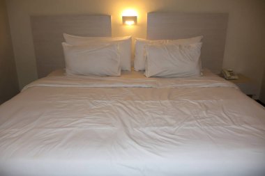 Üzerinde hafif ahşap bir yatak başlığı ve sıcak, düşük yoğunluklu gece lambası bulunan, gevrek beyaz çarşafları ve birden fazla yastığı olan rahat, düzenli bir otel yatağı..