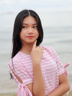 Uzun siyah saçlı, pembe çizgili elbiseli, eli çenesine kadar açık havada poz veren düşünceli bir Asyalı kadının yakın plan portresi..