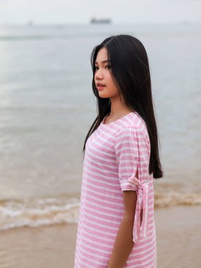 Uzun siyah saçlı, pembe çizgili bir elbisesi olan genç bir Asyalı kadının, sahilde düşünceli bir ifadeyle okyanusa doğru bakan bir yan görüntüsü..