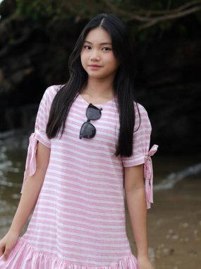 Pembe ve beyaz çizgili uzun siyah saçlı genç Asyalı kadın. Yakasına güneş gözlüğü takıyor. Açık havada, plaj geçmişi bulanık..