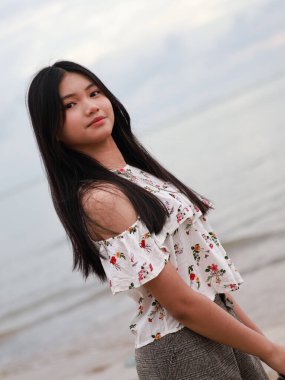 Balikpapan-Endonezya 24 Eylül 2025 güzel, uzun siyah saçlı, bulutlu bir sahilde duran, omuzlarında beyaz çiçekli bir elbise olan genç bir Asyalı kadının portresi..
