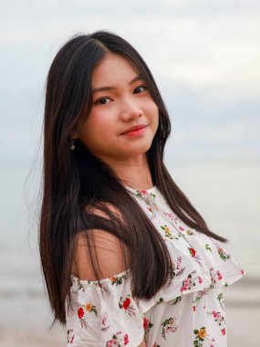 Balikpapan-Endonezya 24 Eylül 2025 Deniz kenarında duran uzun siyah saçlı güzel bir Asyalı kadının yakın plan portresi. Yumuşak gülümsüyor ve yaz tatili için beyaz bir çiçekli bluz giyiyor..