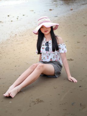 Balikpapan-Endonezya 24 Eylül 2025 Kumlu kumlu bir sahilde oturan uzun siyah saçlı, çiçekli omuz üstü ve geniş kenarlı güneş şapkası takan, açık havada geçen yaz tatilinin tadını çıkaran genç Asyalı kadın..