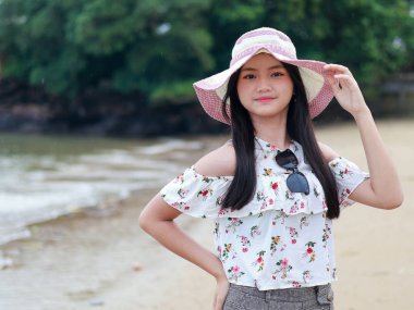 Balikpapan-Endonezya 24 Eylül 2025 Çiçekli, omuzsuz, güneş şapkalı tatlı Asyalı kız, sahilde poz veriyor..