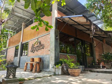 Gorontalo-Endonezya 20 Ekim 2025 Kırsal bir otel ya da resepsiyon binasının dışı kırmızı tuğlalı, ahşap panelli, eğri büğrü metal çatılı, tropikal bitkiler ve güneşli bir ağacın altında ahşap bir bank.