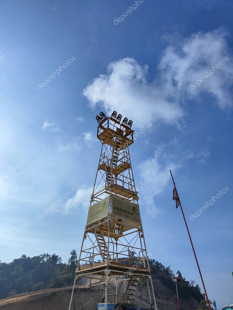 Gorontalo-Indonesia Oct 21, 2025 Low-angle view of a tall, yellow steel lighting tower at a mining or construction site, featuring the 'Merdeka' logo, set against a bright blue sky with clouds.
