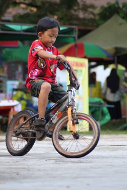 Balikpapan-Endonezya 25 Ekim 2025 Kırmızı tişörtlü genç bir Asyalı panayır ya da karnaval alanından geçerek çamurlu beton bir yüzey üzerinde bisiklet sürüyor..