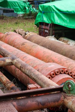 Büyük, yoğun paslanmış hidrolik silindirler ve terk edilmiş çelik borular, yeşil bir muşamba depolama alanının yakınında şiddetli yıpranma ve aşınma gösteriyor..