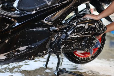 Parlak siyah scooter gövde ve arka tekerlek yıkayan, kalın beyaz sabun köpükleriyle kaplı, araç bakım ve bakım işlemini vurgulayan kişi.