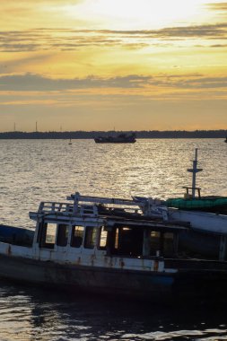 Ön planda eski bir ahşap tekne (balıkçı teknesi) su üzerinde yüzüyor, uzak ufukta daha büyük bir gemi silueti ile altın gökyüzü ve güneş ışığı yansıması çelişiyor.