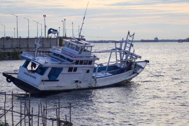 Geleneksel bir Endonezya balıkçı teknesi. Beton iskelenin yanında demirlemiş. Kargo gemileri engin ve bulutlu bir gökyüzünün altında, alacakaranlıkta görülebiliyor..