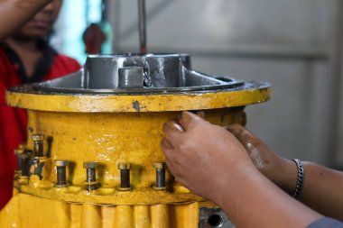 Bir tamirci, önemli bir endüstriyel bakım süreci sırasında flanş kapağını açıkta kalan cıvataların üzerine hizalayarak büyük, sarı bir vites düzenliyor..