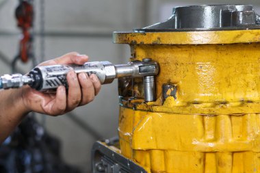 Bir tamirci, bir atölyede gerekli tamir ve montaj çalışmalarını gösteren büyük, ağır sarı bir vites ya da son sürüş ünitesinin sürgüsünü sıkmak için soket anahtarı kullanır..