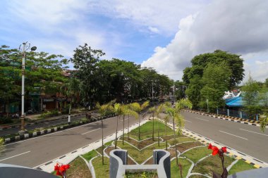 Bereketli yeşil ağaçlar, parlak bir gökyüzünün altında geniş ve sessiz bir şehir yolu boyunca uzanır. İçinde küçük palmiyeler ve kırmızı çiçeklerle dekoratif bir medyan bulunur..