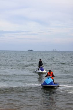 16 Kasım 2025 'te Balikpapan-Endonezya' da iki adam, bulutlu bir gökyüzünün altında, su sporları etkinliği için can yeleği giymiş, sakin deniz kıyısına doğru şahsi su araçları (jet ski) sürüyor..