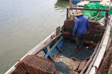 Küçük ahşap bir teknede hasır şapka giyen bir balıkçının yüksek açılı görüntüsü. Etrafı paslı metal yengeç tuzakları ve halat sargılarıyla çevrili..
