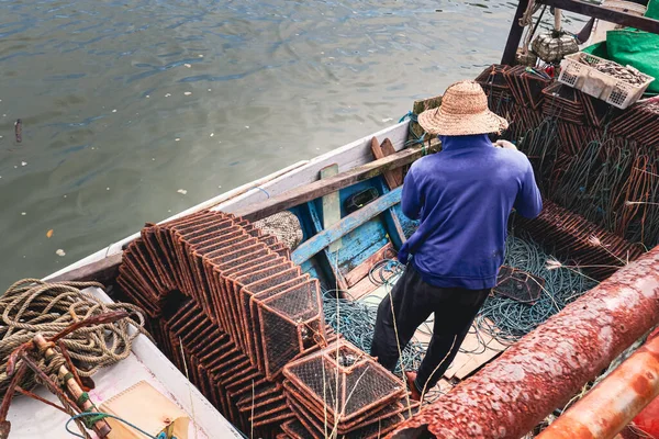 Küçük ahşap bir teknede hasır şapka giyen bir balıkçının yüksek açılı görüntüsü. Etrafı paslı metal yengeç tuzakları ve halat sargılarıyla çevrili..