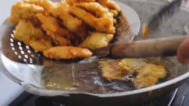 Close-up of golden-brown battered  fried Tempeh being deep-fried in hot oil within a metal wok, with silver tongs used to turn the crispy pieces. Traditional cooking process.