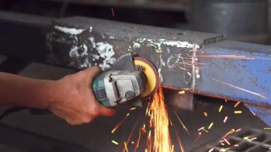 Bir işçinin elinin portatif bir açı öğütücü kullanırken yakın plan çekimi ağır bir çelik kirişi keserken parlak turuncu ve sarı kıvılcımlar saçar..