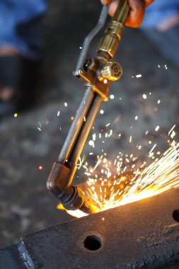 Bir el, ağır çelik kirişi karanlık bir atölyede ısıtıp keserken yoğun alev ve parlak kıvılcımlar üreten bir meşaleyi tutuyor..