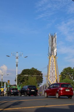 Balikpapan-Endonezya 29 Ocak 2026 Modern şehir anıtı mavi gökyüzü altında geleneksel desenlerle birlikte. Aydınlık bir günde asfalt yolda motosiklet süren motosikletçiler.