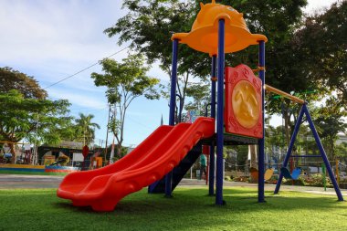 Renkli çocuklar, açık mavi gökyüzünün altındaki ağaçlarla çevrili yeşil çimlerin üzerinde kırmızı çift slaytlar ve turuncu çatılarla kayıyorlar.