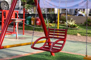 Parlak gün ışığı altında yeşil suni çimleri ve renkli park geçmişi olan bir çocuk parkında zincirli kırmızı metal sallanan koltuk..
