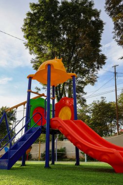 Renkli çocuklar, açık mavi gökyüzünün altındaki ağaçlarla çevrili yeşil çimlerin üzerinde kırmızı çift slaytlar ve turuncu çatılarla kayıyorlar.