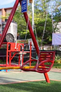 Parlak gün ışığı altında yeşil suni çimleri ve renkli park geçmişi olan bir çocuk parkında zincirli kırmızı metal sallanan koltuk..