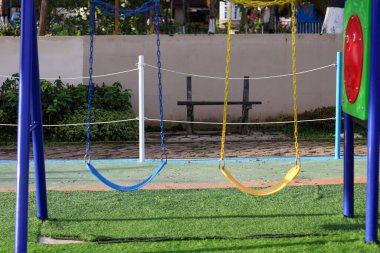 Mavi ve sarı çocuklar yeşil bir çimenlikte salıncakta sallanıyor açık mavi gökyüzünün altında renkli oyun yapıları ve yeşil ağaçlar var..