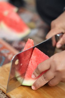 Ahşap bir tahtada olgun kırmızı bir karpuz dilimini kesmek için büyük metal bir satır kullanarak ellerinizi kapatın. Mutfak ortamında taze organik meyve atıştırmalıkları hazırlayın..