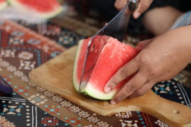 Ahşap bir tahtada olgun kırmızı bir karpuz dilimini kesmek için büyük metal bir satır kullanarak ellerinizi kapatın. Mutfak ortamında taze organik meyve atıştırmalıkları hazırlayın..