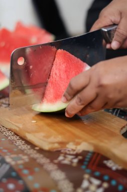 Ahşap bir tahtada olgun kırmızı bir karpuz dilimini kesmek için büyük metal bir satır kullanarak ellerinizi kapatın. Mutfak ortamında taze organik meyve atıştırmalıkları hazırlayın..
