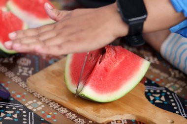 Ahşap bir tahtada olgun kırmızı bir karpuz dilimini kesmek için büyük metal bir satır kullanarak ellerinizi kapatın. Mutfak ortamında taze organik meyve atıştırmalıkları hazırlayın..