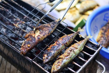 Bir insan gün boyunca dışarıda kömür ocağında taze balık ızgara yapar, maşayla onları dikkatlice çevirerek mükemmel dumanlı, kömürleşmiş bir doku ve lezzetli bir lezzet elde eder..