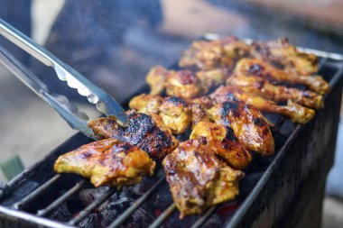 Bir şefin mangal mangalında ızgara terbiye edilmiş tavuk parçaları pişirirken yakın plan çekimi, kömür ızgarasının üzerinde metal maşalar kullanarak eti mükemmel dumanlı, kömürleşmiş bir doku ve lezzetli bir lezzet elde etmek için çevirmesi..