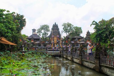 Bali 'nin Ubud kentindeki Saraswati Tapınağı' nın geniş açılı manzarası. Turistler bir nilüfer havuzunun üzerinden geçiyorlar. Süslü Hindu kapısı, tropikal ortam, keskin odak. Seyahat, kültür ve ruhani temalar için ideal.