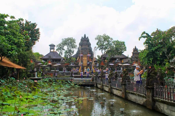 Bali 'nin Ubud kentindeki Saraswati Tapınağı' nın geniş açılı manzarası. Turistler bir nilüfer havuzunun üzerinden geçiyorlar. Süslü Hindu kapısı, tropikal ortam, keskin odak. Seyahat, kültür ve ruhani temalar için ideal.