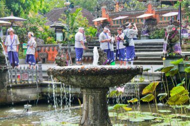 Geleneksel giyinmiş turistler Bali 'nin Ubud kentindeki Pura Saraswati tapınağını ziyaret ediyorlar. Nilüfer havuzunda bir taş çeşme, süslü tapınak kapısı ve parlak bir gökyüzünün altında binalar. 11 Eylül 2024-Endonezya.