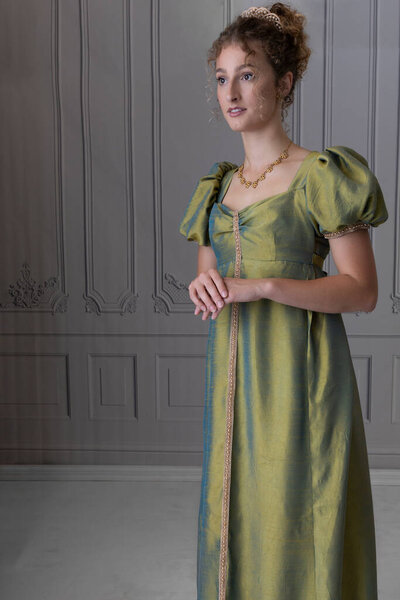 A young Regency woman wearing a green shot silk dress and standing alone in a room with paneled walls 