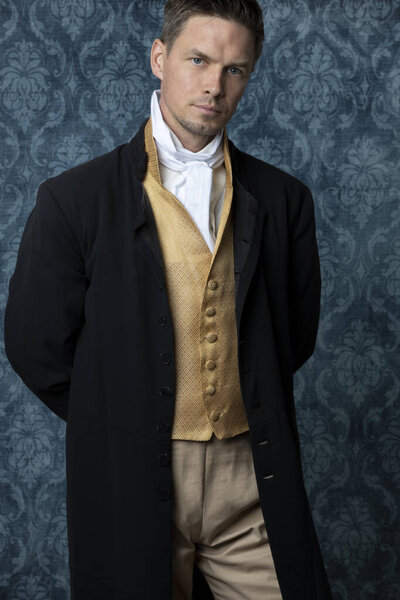 A handsome Regency man wearing a gold waistcoat, breeches, and a black jacket and standing in a room with blue wallpaper and a wooden floor. 