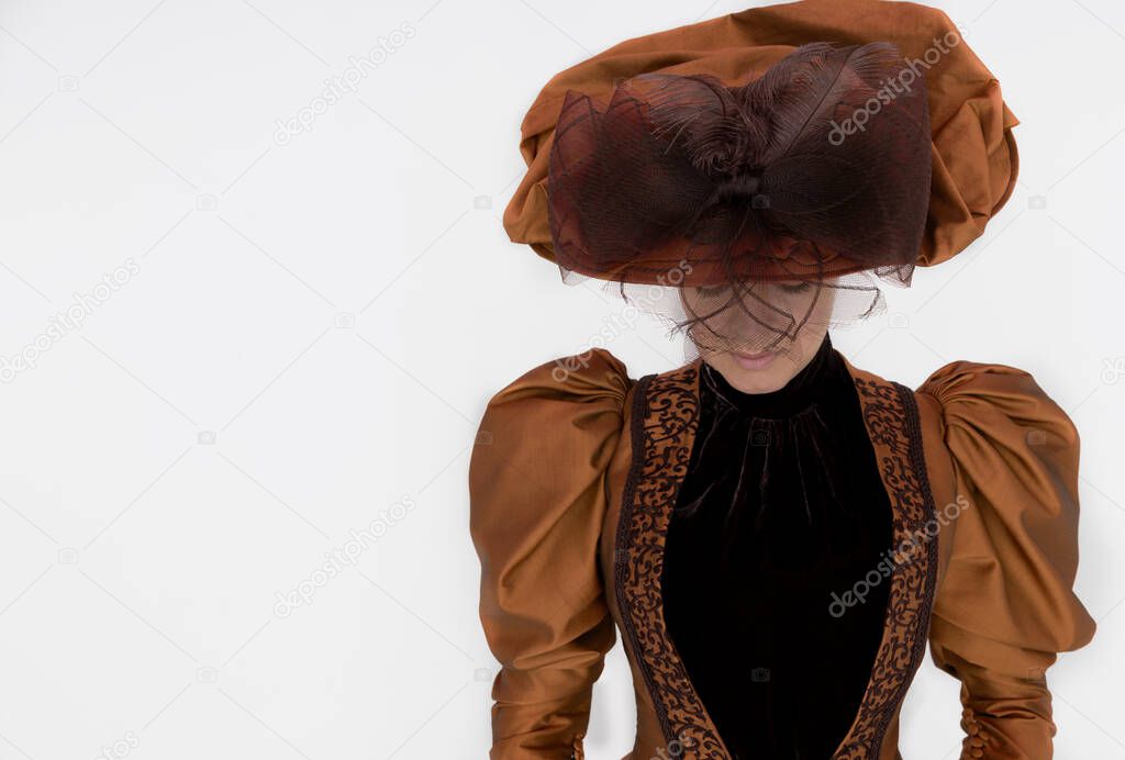 A Victorian woman wearing a bronze and brown silk ensemble and a large hat and posing in front of a white backdrop