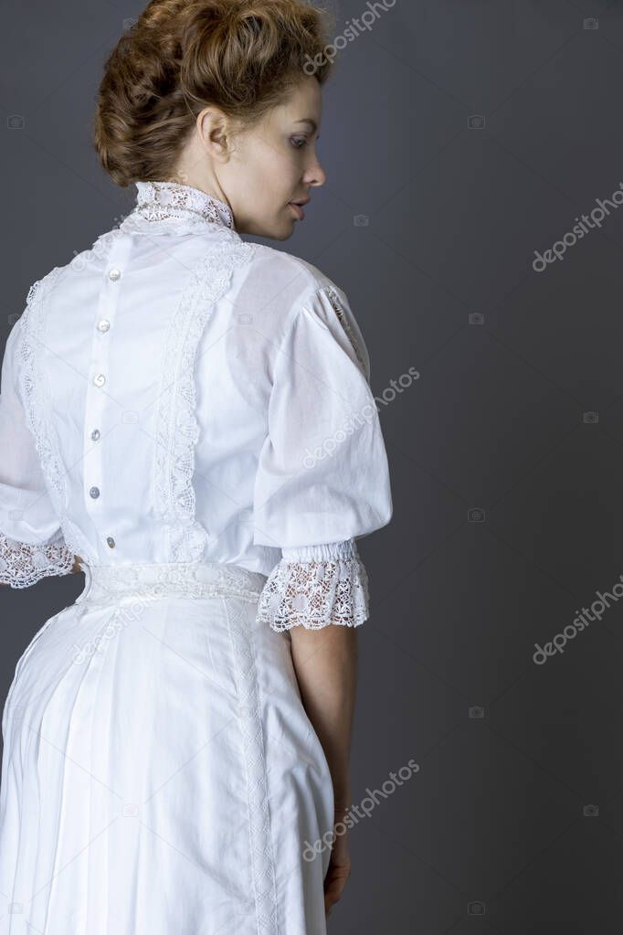 A Edwardian woman wearing a white lace blouse and skirt with a long pearl necklace against a grey studio backdrop 