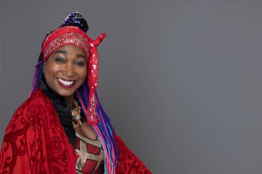 Parlak renkli kıyafetler giyen ve gri bir stüdyo arkaplanına başörtüsü takan gülümseyen bir kadın.