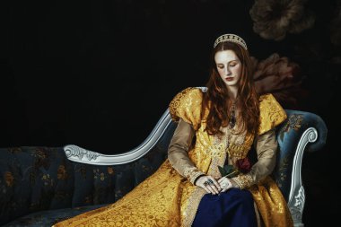 Altın sarısı bir Rönesans elbisesi giyen, kabarık kollu, nakışlı bir koltukta oturan kızıl saçlı bir kadın.