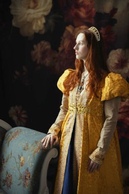 Kizil saçli bir kadin, Rönesans elbisesi giyiyor, kabarık kollu, tek basina duruyor.