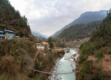 Nepal dağlarında nehir kıyısında küçük bir dağ kasabası ve sallanan bir köprü.