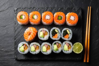 Siyah taşta servis edilen somon ve avokadoyla birlikte yaratıcı suşi ruloları. Taze limon dilimi ve koyu arka planda ahşap yemek çubukları içerir.