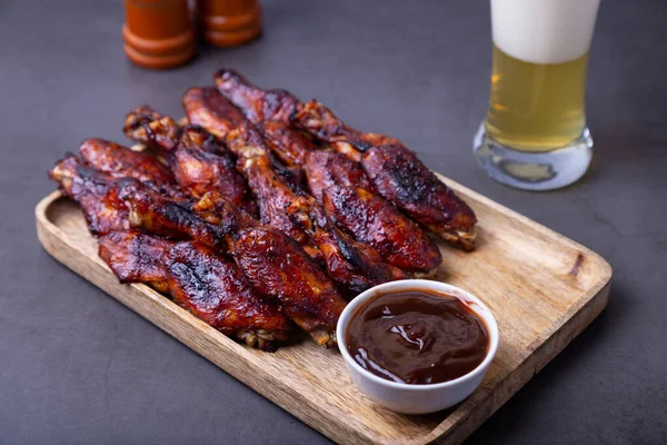 Ahşap bir tahtanın üzerinde kızarmış tavuk kanadı, barbekü sosu ve bir bardak bira. Siyah arkaplan, yakın plan.